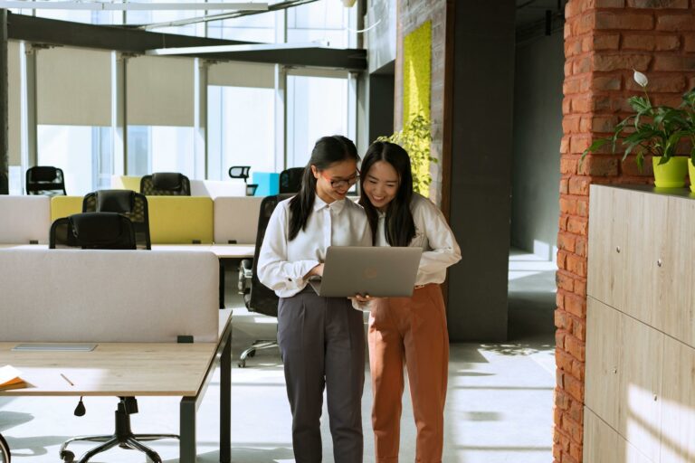 Two women stand together in a bright office space, collaborating on a laptop.